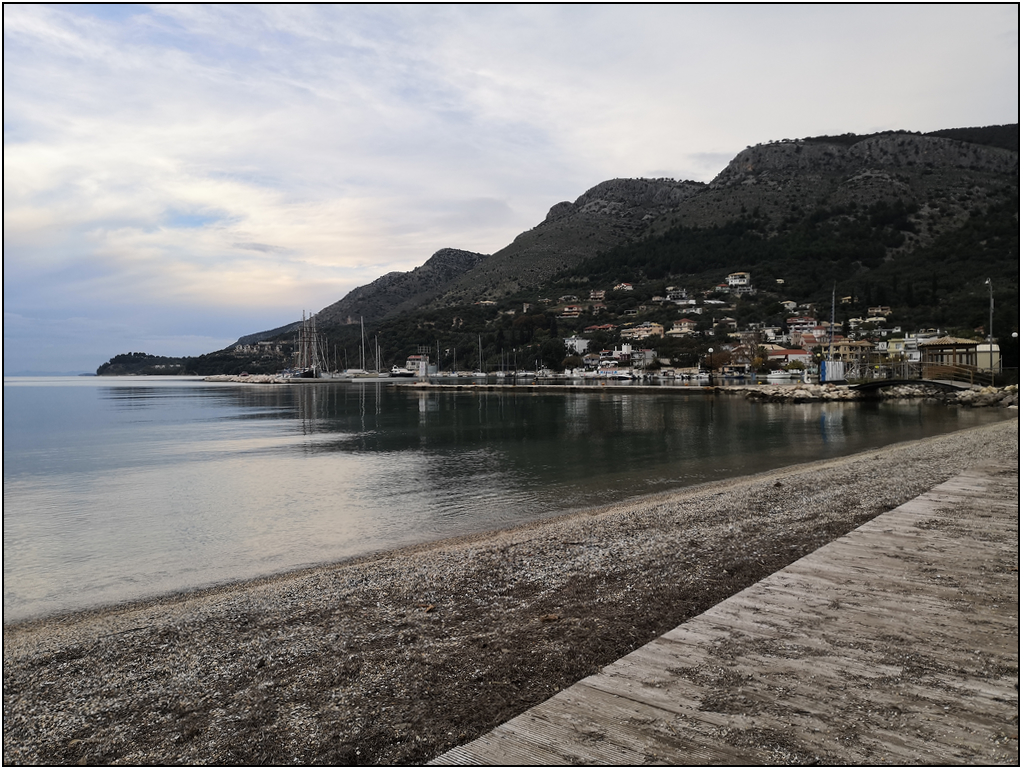 Photo de la plage de Plataria. AU second plan le petit port et les montagnes. Ciel couvert