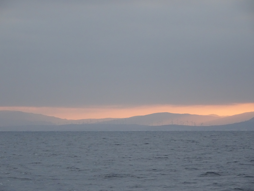 Mer et ciel sont gris, la côte se découpe dans un ciel orangé. Les éoliennes sont à peine visible dans la brume